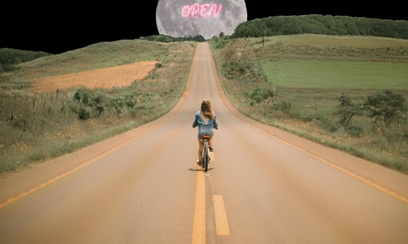 Eine Person fährt mit dem Fahrrad auf einer leeren Landstraße in Richtung eines großen Mondes, auf dem das Wort 'OPEN' leuchtet.