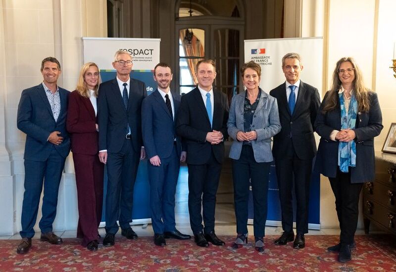 CEO Talk: Innovation als Schlüssel zur Transformation / Philia Kleemann Eine Gruppe von acht Personen steht lächelnd in einem eleganten Raum, gekleidet in formellen Anzügen. Im Hintergrund sind zwei Banner zu sehen, eines mit der Aufschrift 'Ambassade de France en Autriche'. Der Boden ist mit einem roten Teppich bedeckt,